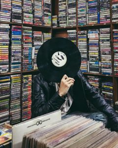 A man in a leather jacket sits holding a record over his face. In the foreground are bins full of records, the walls behind him are floor to ceiling cassette racks that are full. PRIR. Pocket Rock It Radio. Listen. Even More. Cave.